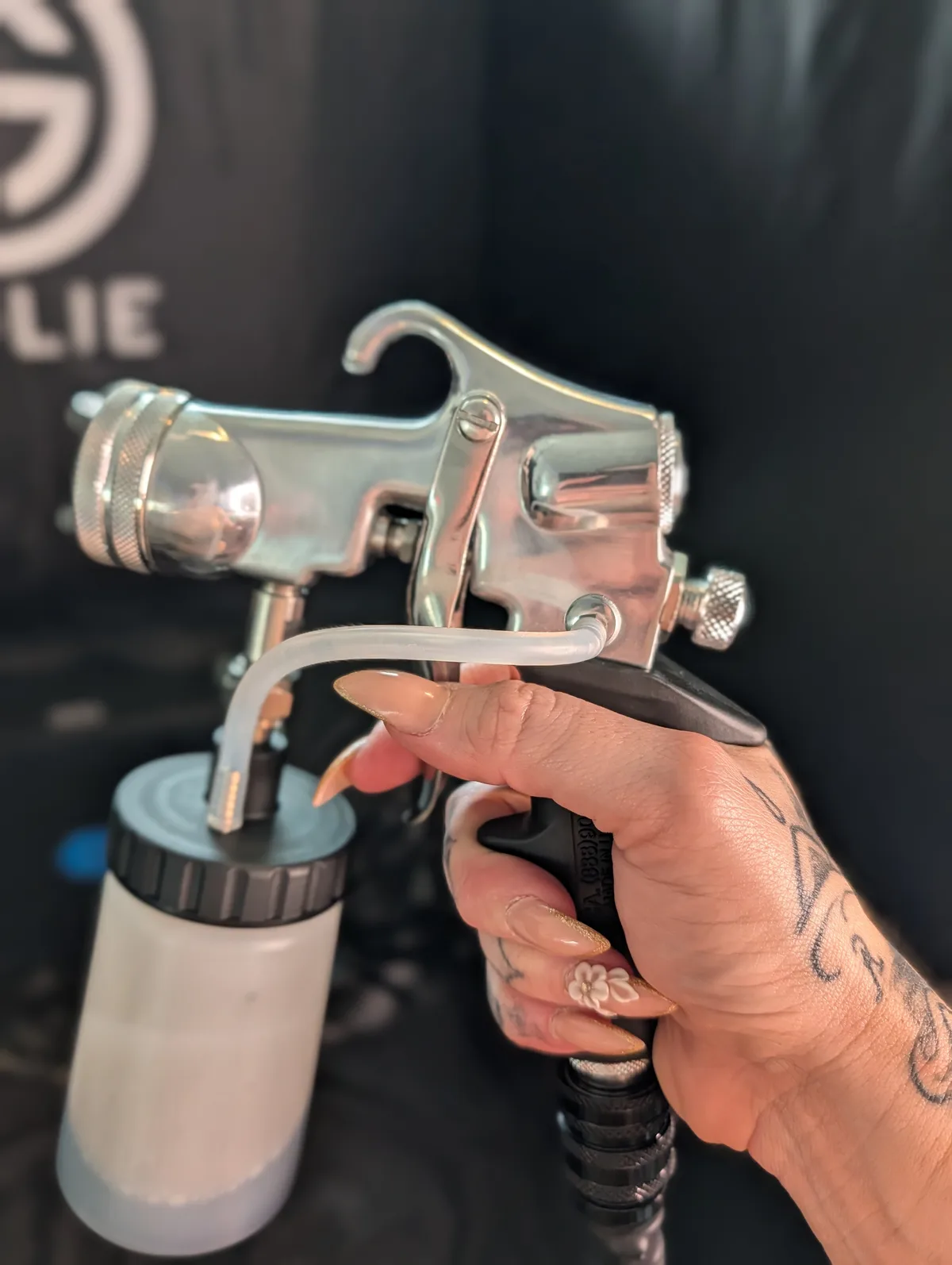 Glowtoure spray tan artist holding the airbrush gun, custom-mixed solution ready, against the branded Glowtoure backdrop